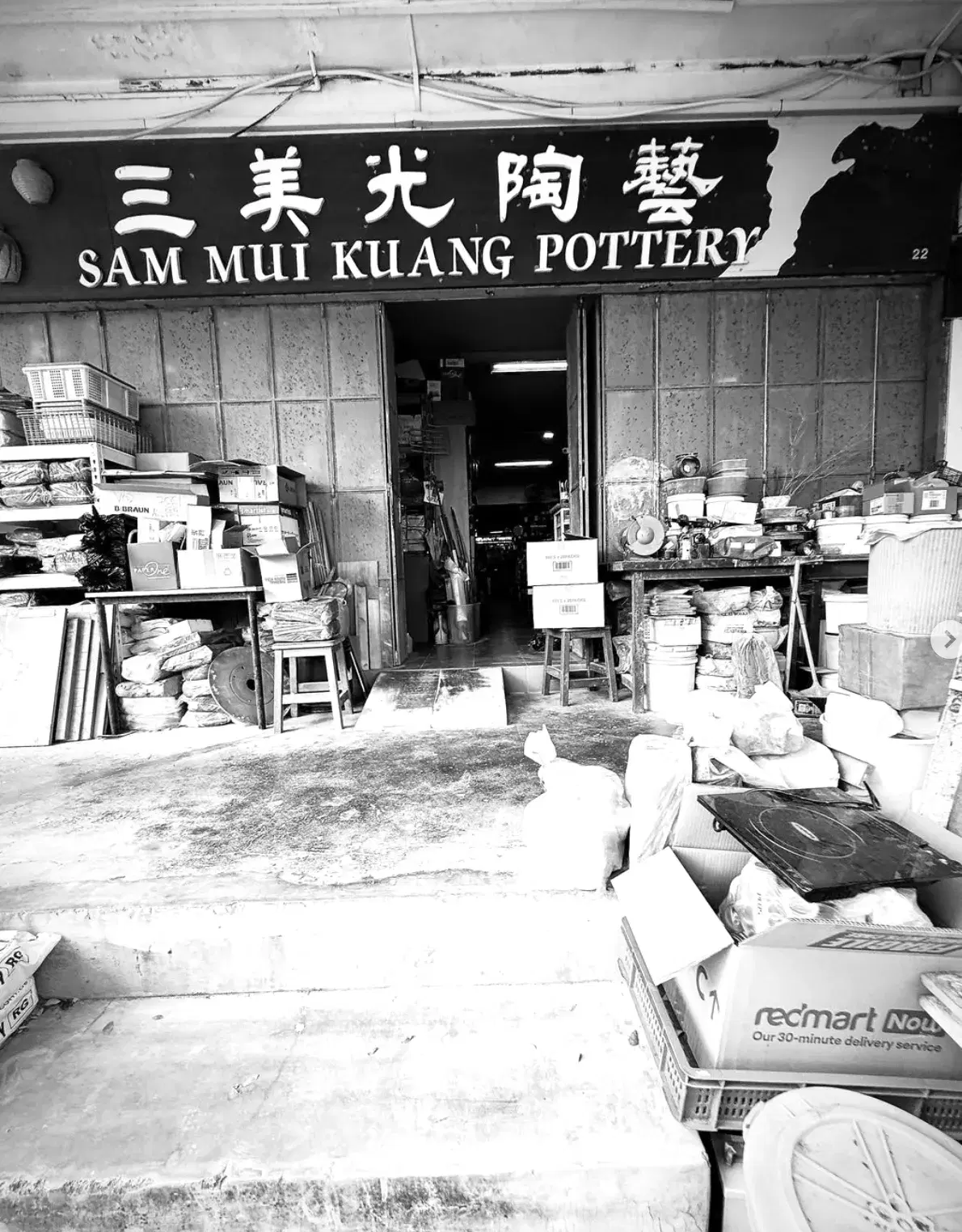 Black-and-white photo of the Sam Mui Kuang Pottery shopfront at 22 Jalan Kelulut, with its bilingual signboard 三美光陶艺 above an open doorway flanked by stacked boxes, buckets and pottery supplies