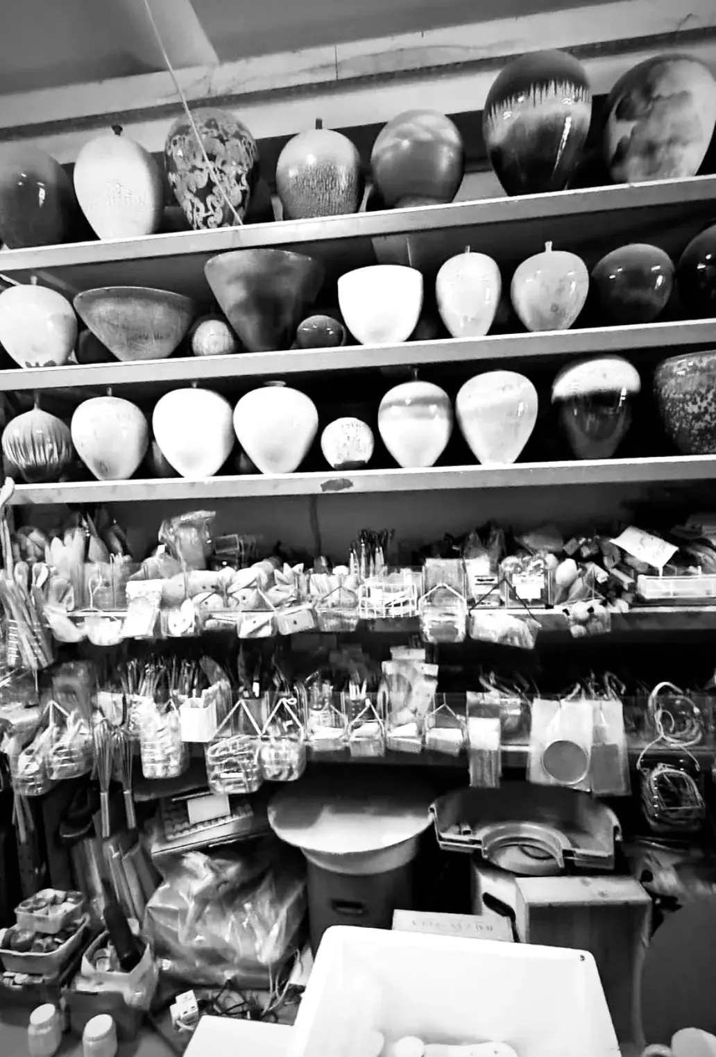 Black-and-white photo of studio shelves lined with rows of crystalline-glazed vases and bowls, with pottery tools and supplies hanging below
