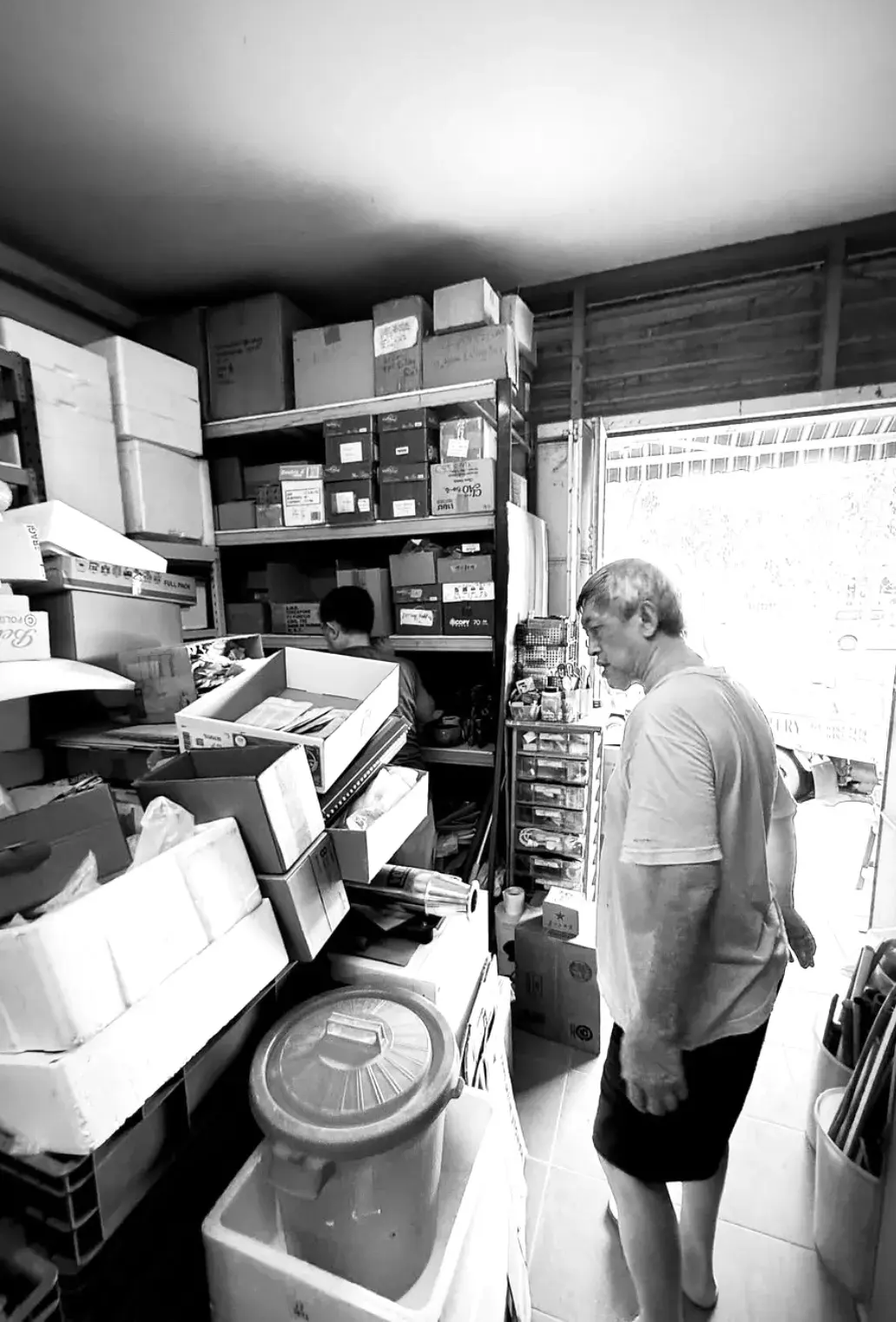 Black-and-white photo of Mr. Chua Soo Kim inside the studio storeroom, surrounded by shelves of boxed supplies, glaze materials and a covered bucket